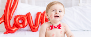 Baby with a red bowtie against a white backdrop and a red balloon spelling the word "love".